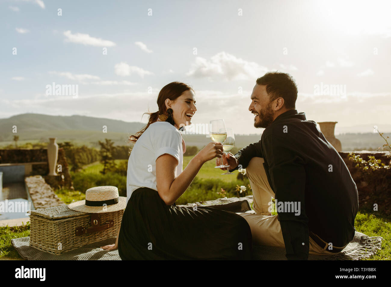 Sorridente giovane seduto in un vino azienda agricola di bicchieri di vino bianco. Giovane su di una data seduta in un vigneto con un cestino da picnic al loro fianco. Foto Stock
