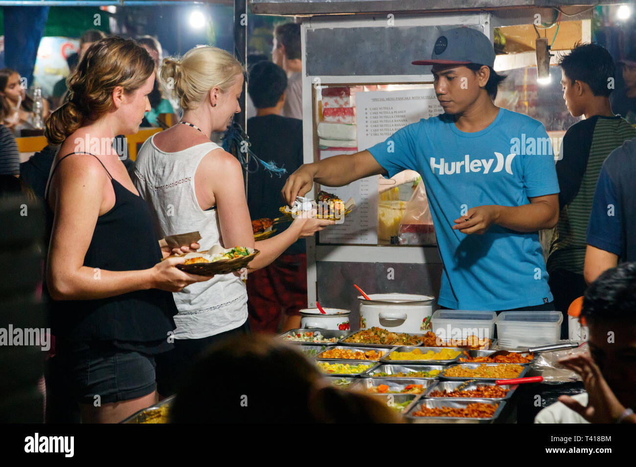 Ragazzo che serve cibo per un turista femminile presso il locale mercato alimentare a notte. Gili Trawangan, West Nusa Tenggara, Indonesia. Foto Stock