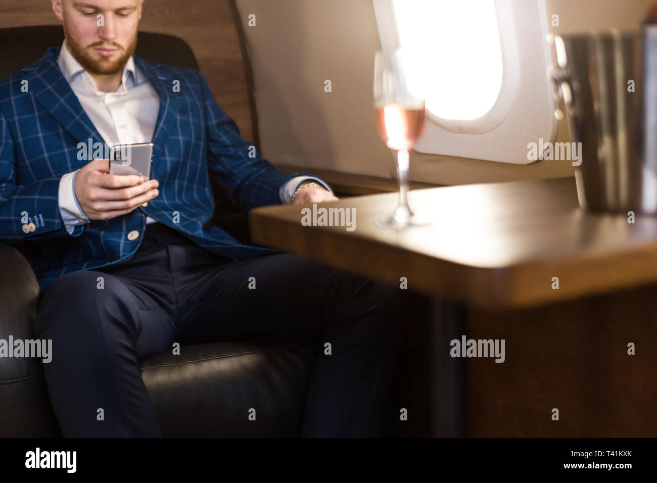 Un giovane imprenditore di successo in un costoso abito si siede sulla poltrona di un jet privato con un bicchiere di champagne in mano e guarda fuori dalla finestra. Foto Stock