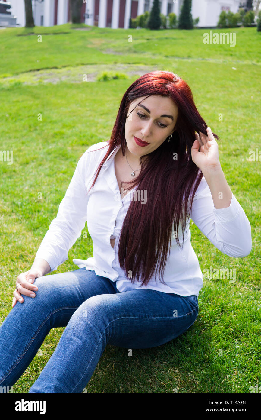 Giovane donna in maglia bianca e jeans blu seduti su un prato verde sul  parco della città prato Foto stock - Alamy
