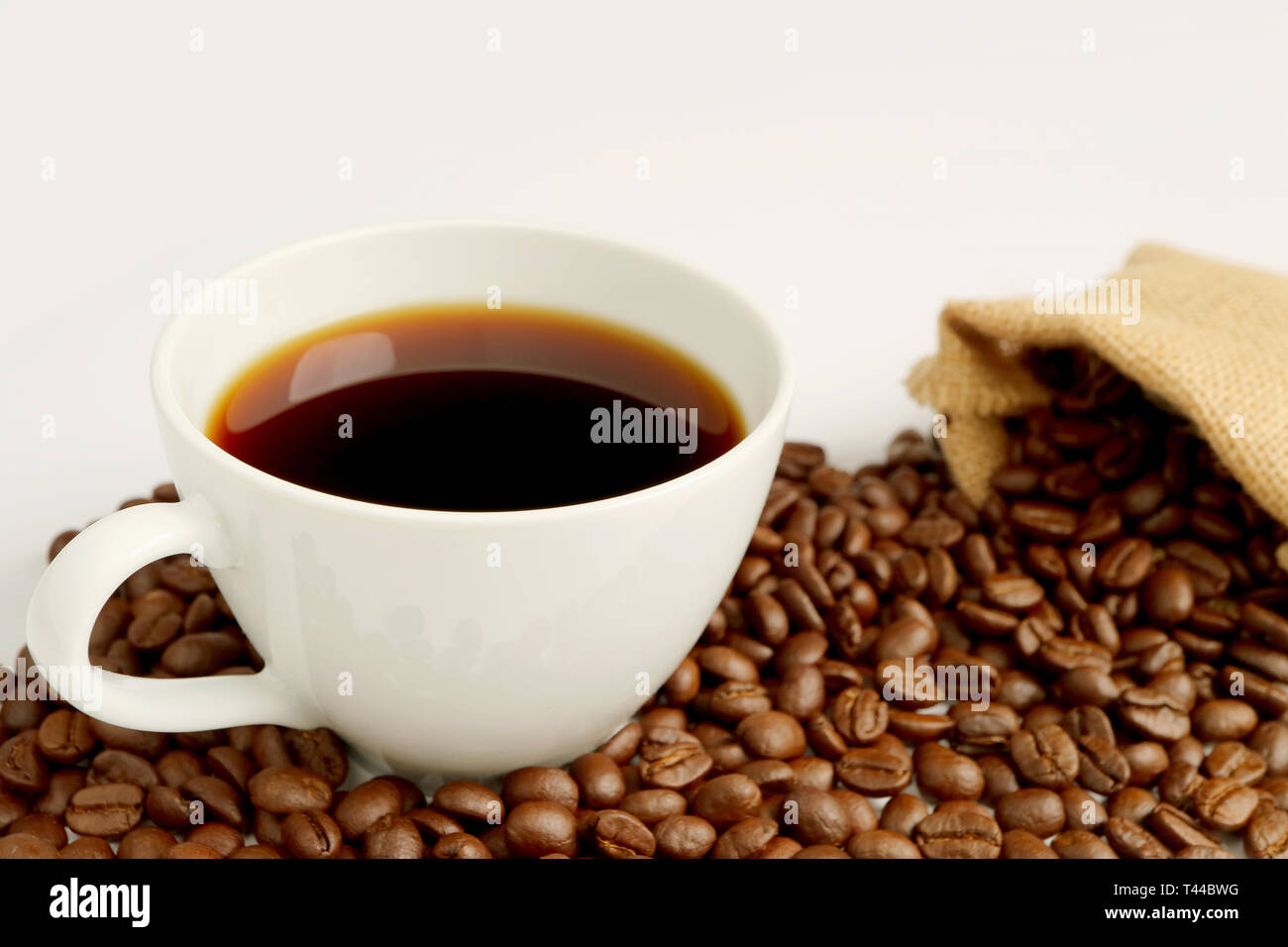 Una tazza di caffè nero con la pila di caffè torrefatto in grani sparsi dal sacchetto di tela su sfondo bianco Foto Stock