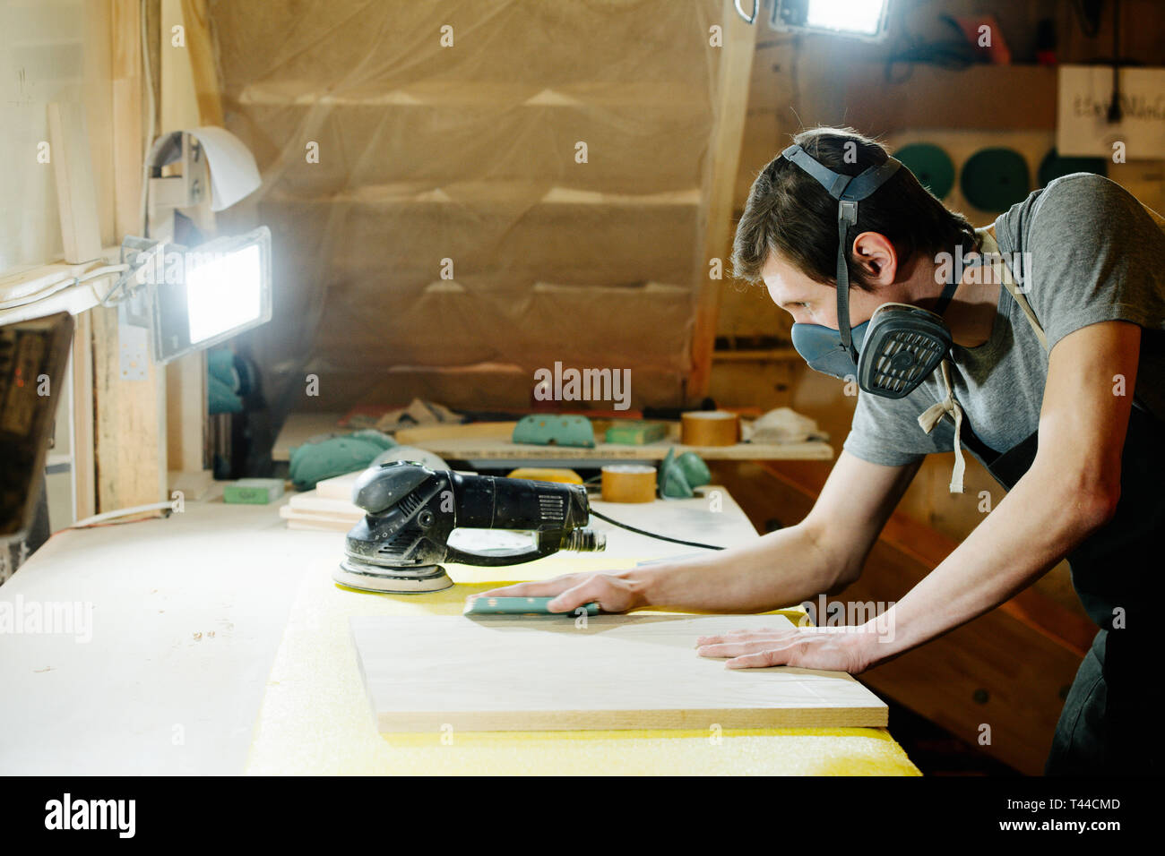 Ritratto di lavoro slim bello di mezza età brunet carpenter nel respiratore. Egli sta rendendo mobili su ordine in un workshop. Levigatura del pannello di legno surf Foto Stock