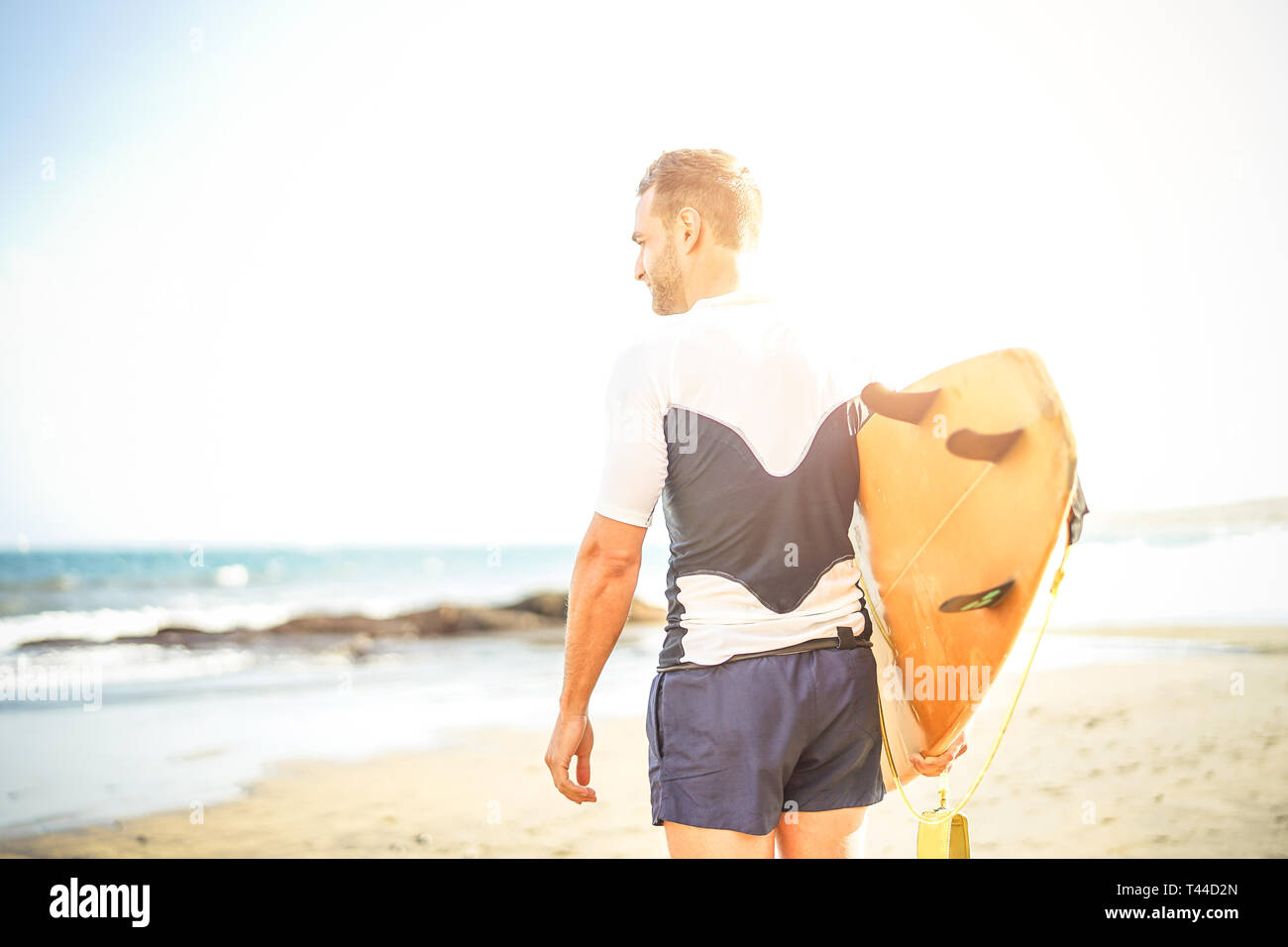 Giovani surfer tenendo la sua tavola da surf guardando le onde per il surf - uomo bello permanente sulla spiaggia al tramonto la formazione per il surf Foto Stock