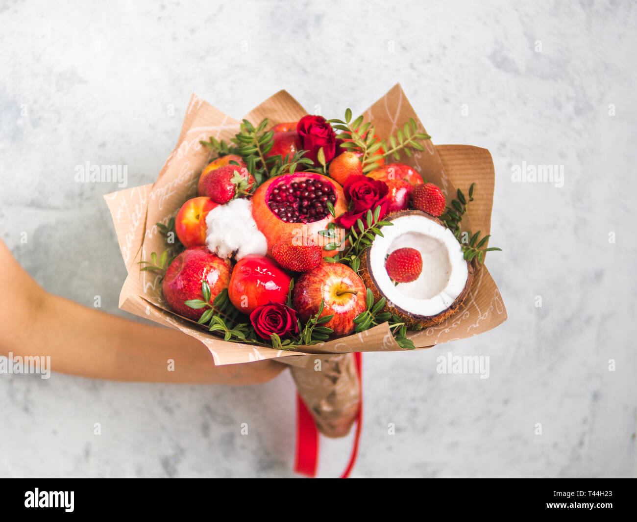 Frutta e bacche bouquet. Mangiare bouquet in mano femmina su sfondo grigio. Melograno, fragola, mele, noci di cocco e le rose fiori, eucalipto. DOF poco profondo. Spazio di copia Foto Stock