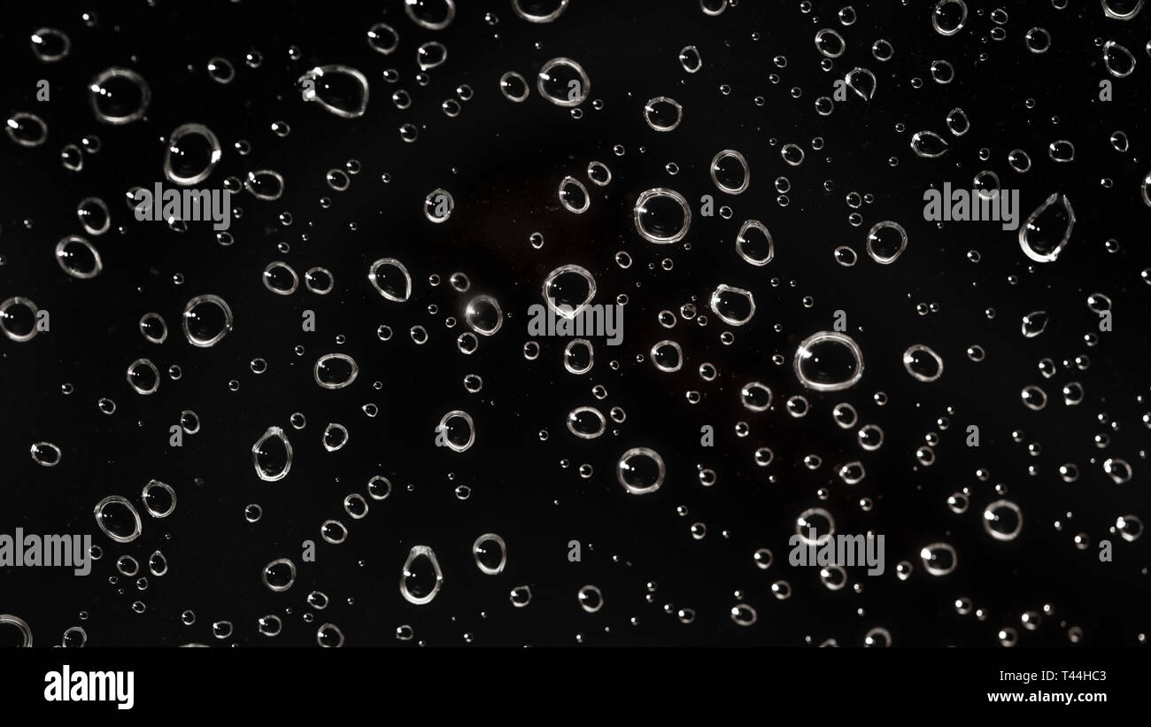 Primo piano - La pioggia di gocce d'acqua dietro il vetro simile a bolle nel liquido nero. Abstract sfondo umido. Foto Stock