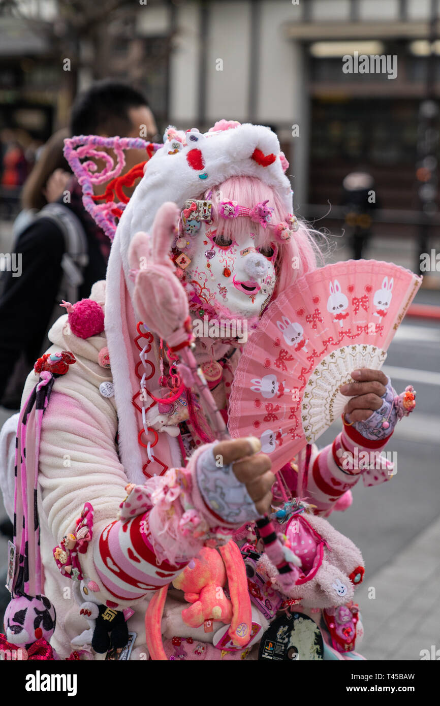 Tokyo, Harajuku è il gioco cos il centro di Tokyo con vari giovani mode in evidenza. Questo uomo, noto come Kadocchi, è considerato come egli unoffical icona di Harajuku. Uomo vestito da clown in rosa e bianco vestito mantenendo la ventola, in posa per il visualizzatore. Contatto visivo. Foto Stock
