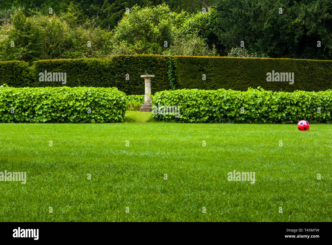 Doppio hedge con Meridiana su un plinto visto in un traferro e con un calcio rosso giacente sul prato, rendendolo una combinazione di formale e informale Foto Stock