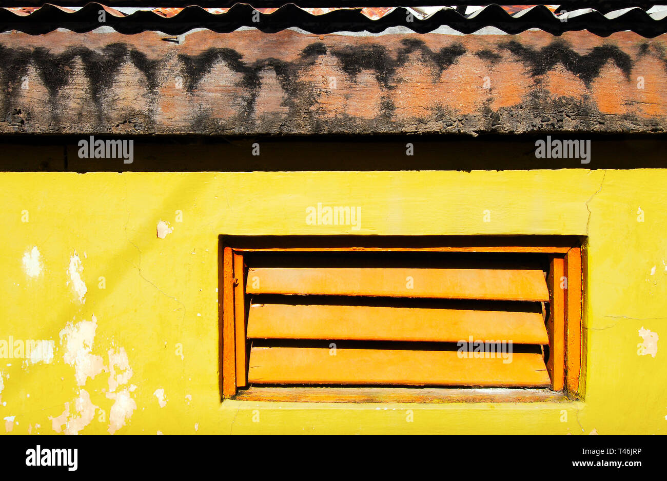 Colore arancio persiane alle finestre in un giallo brillante parete dipinta con ombre scure dal tetto in legno con lamiere grecate Foto Stock