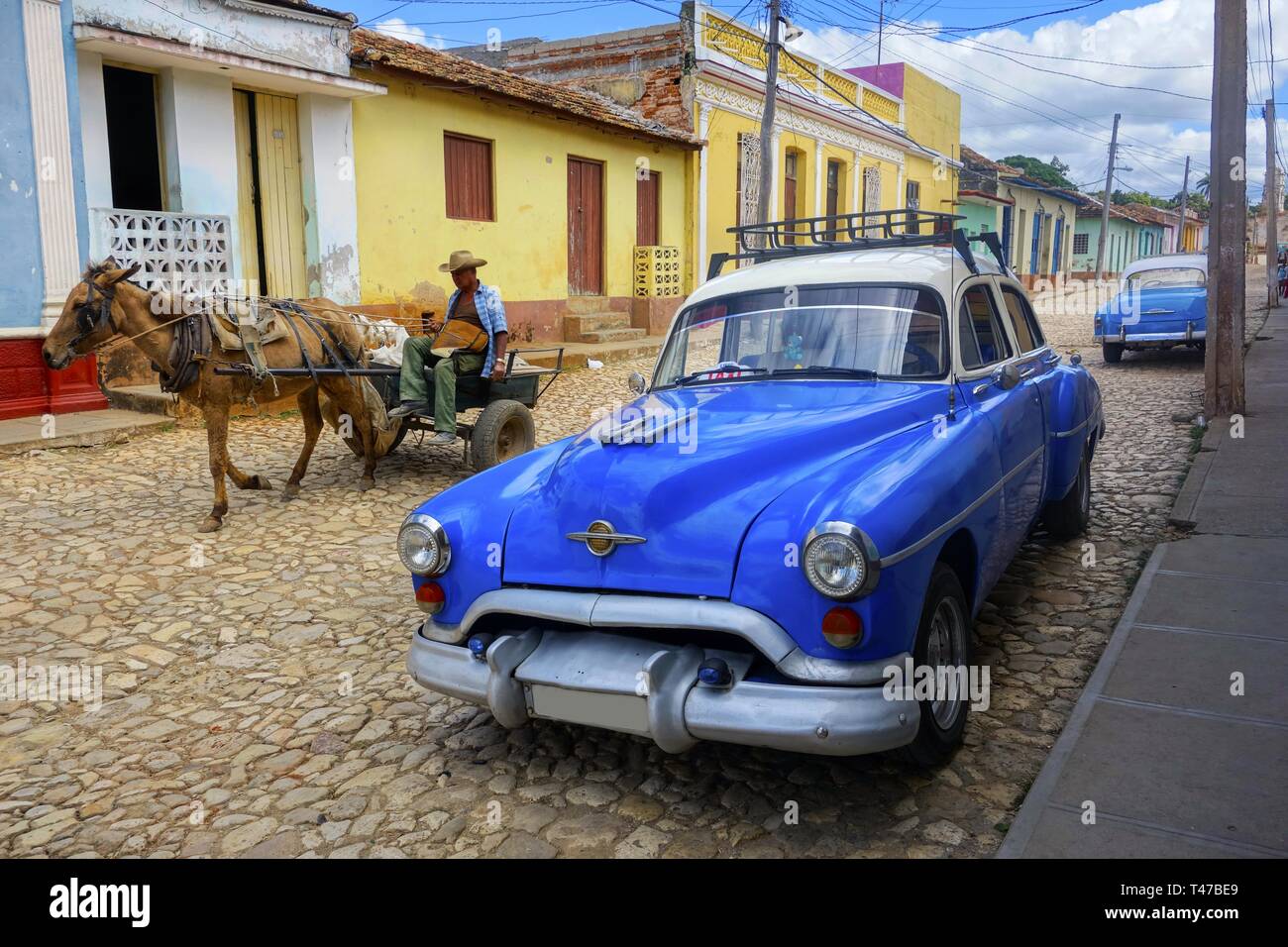Old Man in carrozza a Cavallo e Classic Cuban Taxi Car su Cobblestone Street che mostra modalità di trasporto contrastanti e la tipica vita cittadina di Trinidad, Cuba Foto Stock