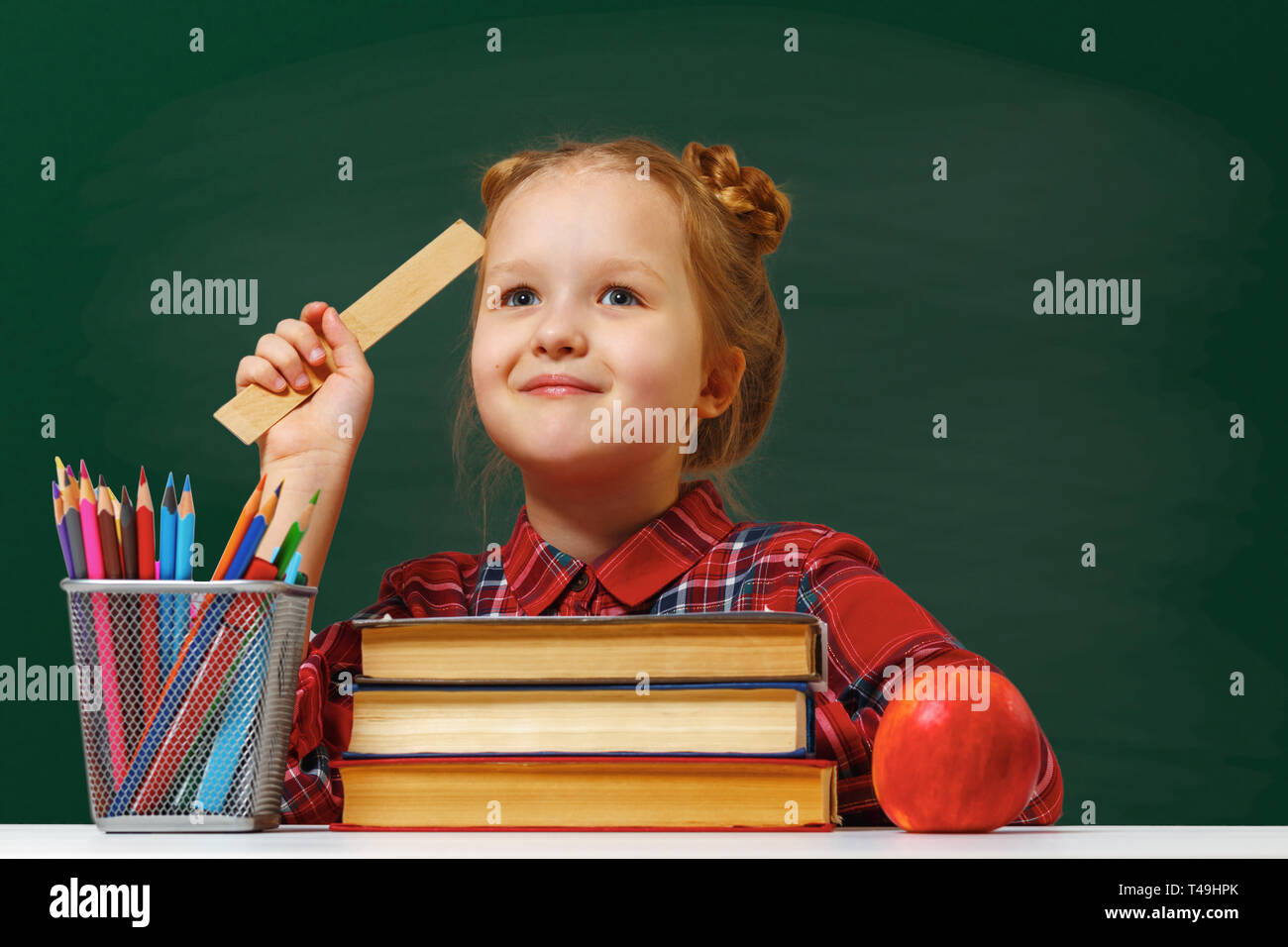 Un simpatico bambina premurosamente si siede a un tavolo. La scolaretta è circondato da libri e matite sullo sfondo di una green school board. Foto Stock