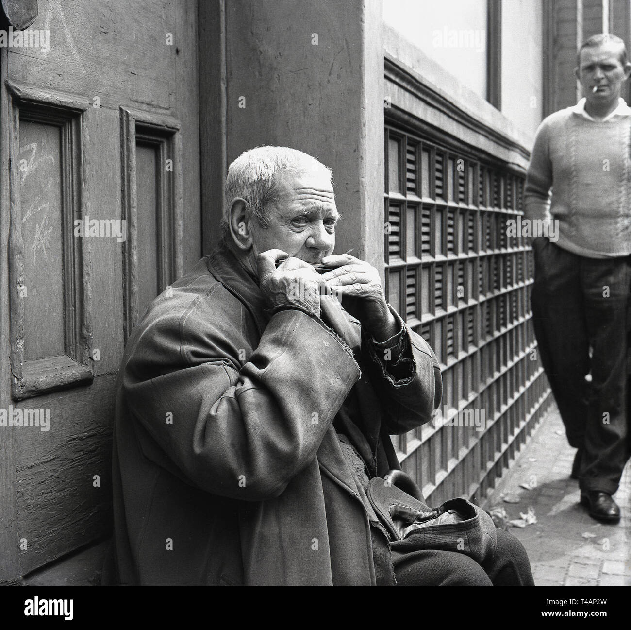 Anni sessanta, storico, un busker, un anziano uomo seduto in una strada porta con la sua calotta aperta nel suo giro, la riproduzione di un'armonica o armonica a bocca da Stepney mercato, East London, Londra, Foto Stock