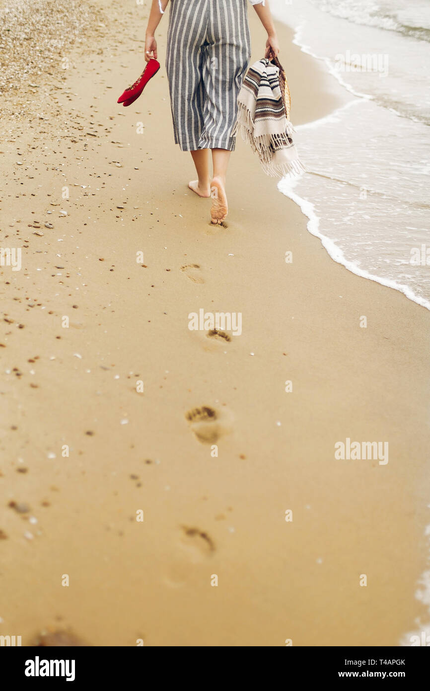 Donna camminare a piedi scalzi sulla spiaggia, vista posteriore delle  gambe. Giovane ragazza rilassante sulla spiaggia di sabbia, a piedi con le  scarpe e la borsa in mani. La vacanza estiva concept
