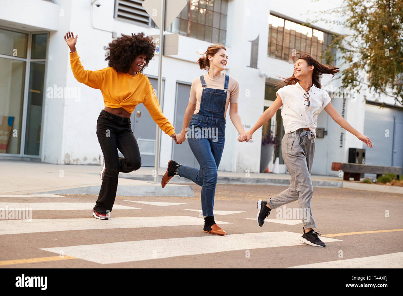 Tre anca millenario fidanzate tenendo le mani e ridendo come essi corrono attraverso un passaggio pedonale Foto Stock