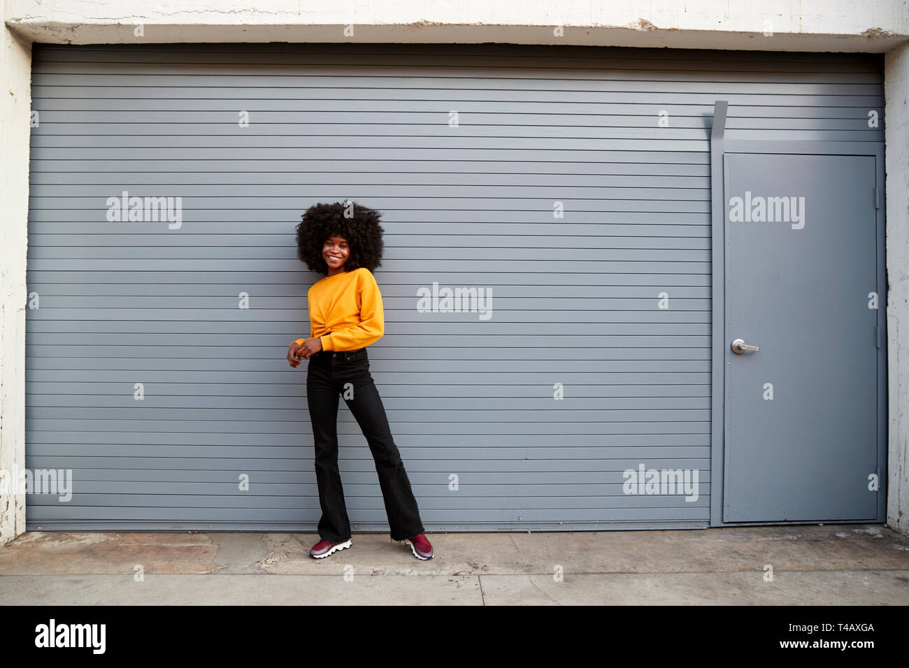 Hip giovane nero donna in piedi contro il grigio a tapparelle di sicurezza sorridente alla telecamera a piena lunghezza Foto Stock