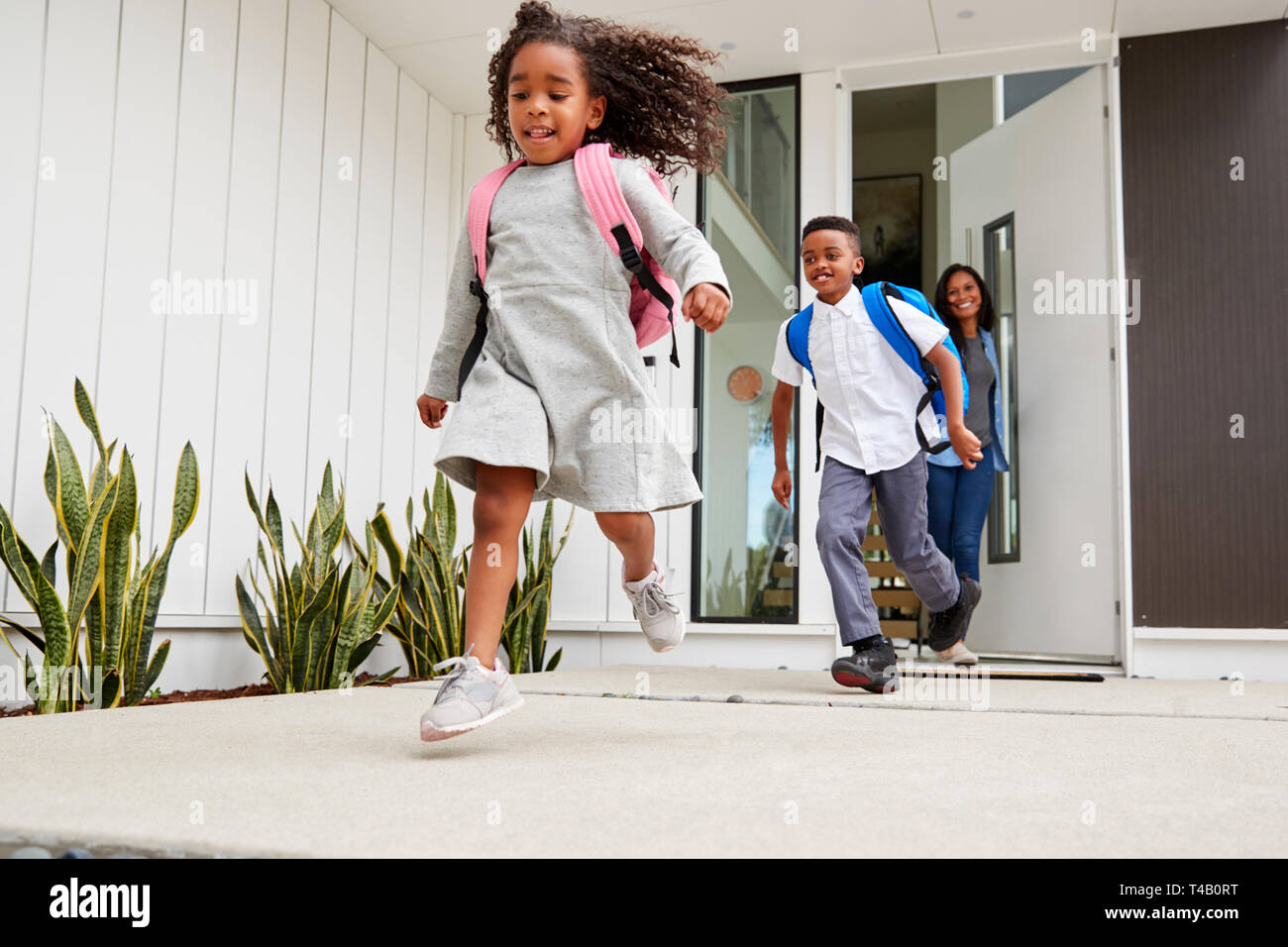 Emozionato i bambini in esecuzione fuori della porta anteriore sulla strada per la scuola guardato da madre Foto Stock