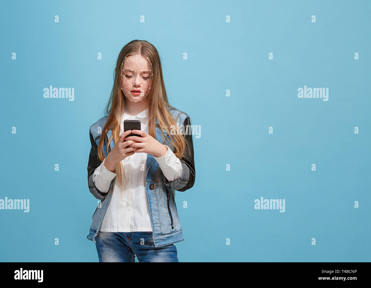 Tempo per nuove conoscenze. Una ragazza con uno smartphone in jeans tuta in piedi su sfondo blu. La tecnologia di riconoscimento facciale su griglia poligonale. Concetto di cyber security, business, lavoro, educazione. Foto Stock