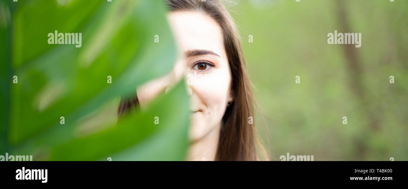 Le bellezze naturali della giovane donna closeup dietro grandi foglie di monstera con sfondo verde nei boschi. La bellezza, la salute e la natura del concetto. Foto Stock