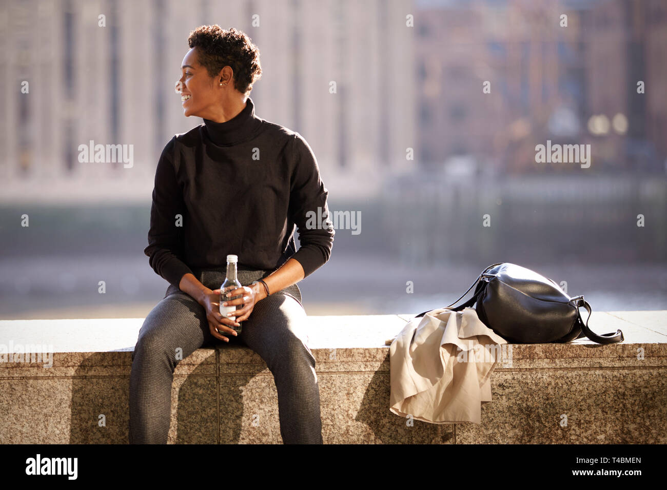 Giovane nero donna che indossa un maglione turtleneck seduti al sole su un terrapieno parete mediante il Fiume Tamigi a Londra, retroilluminato Foto Stock