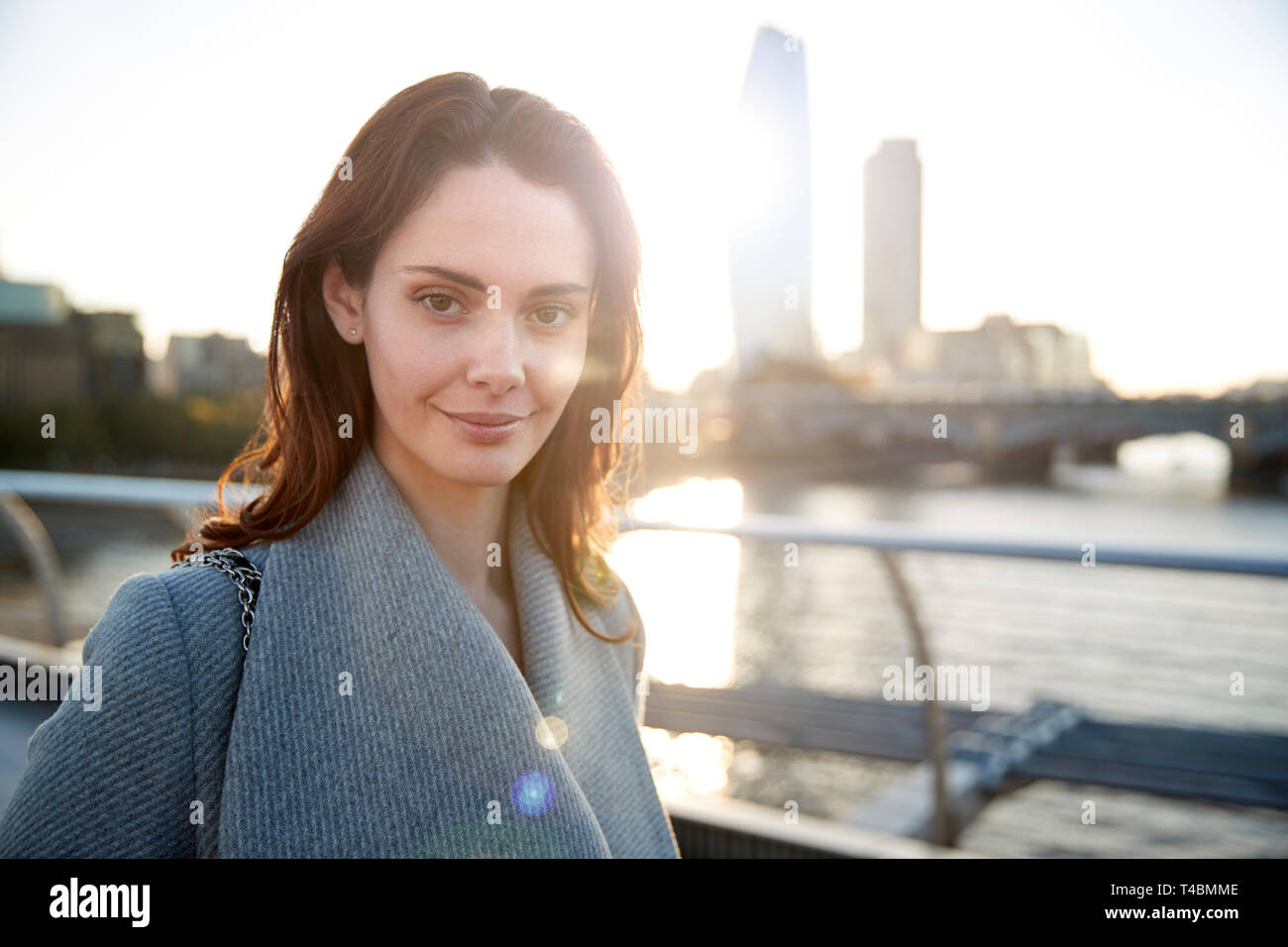 Bianco giovane donna che indossa cappotto grigio in piedi sul Millennium Bridge, Londra, cercando di fotocamera a sorridere Foto Stock