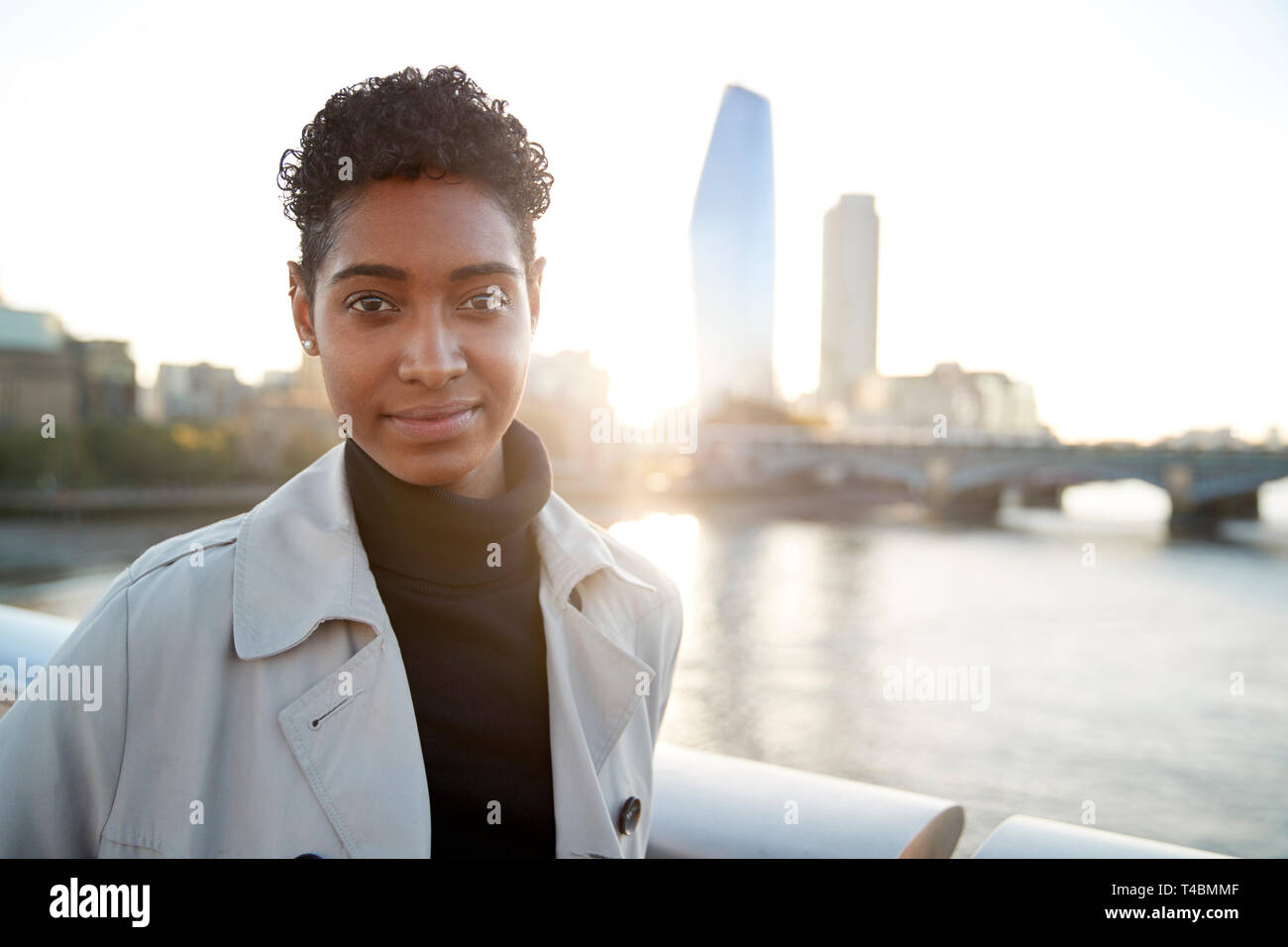 Giovane nero donna che indossa un maglione turtleneck e un mackintosh in piedi sul Millennium Bridge, Londra, cercando di fotocamera a sorridere Foto Stock