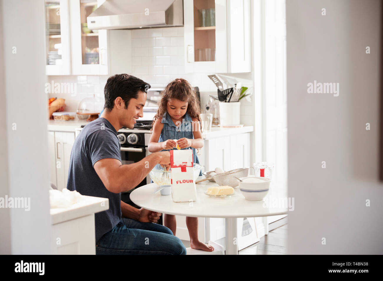 Ragazza giovane permanente al tavolo della cucina la preparazione di miscela per torte con suo padre, visto dalla porta Foto Stock