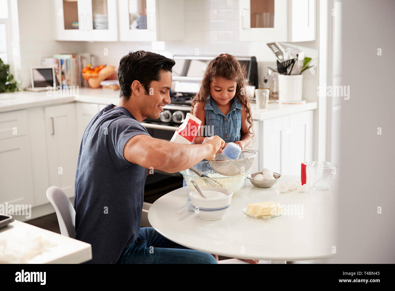 Ragazza giovane permanente al tavolo della cucina la preparazione di miscela per torte con suo padre, visto dalla porta Foto Stock