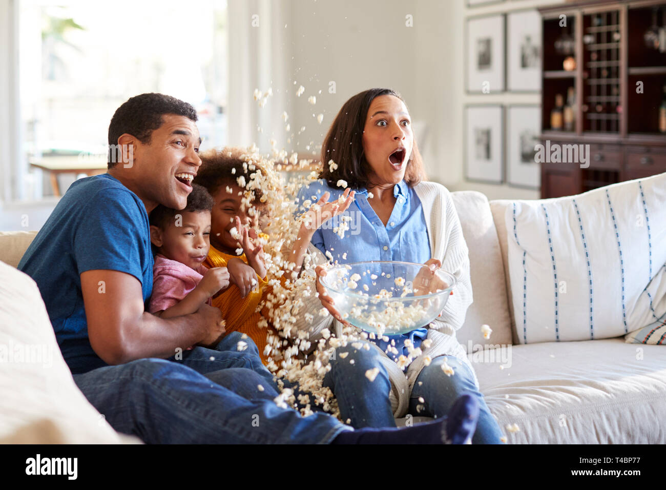 Giovani razza mista famiglia seduti insieme sul divano nel loro salotto guardando un film spaventoso accidentalmente popcorn gettando in aria Foto Stock