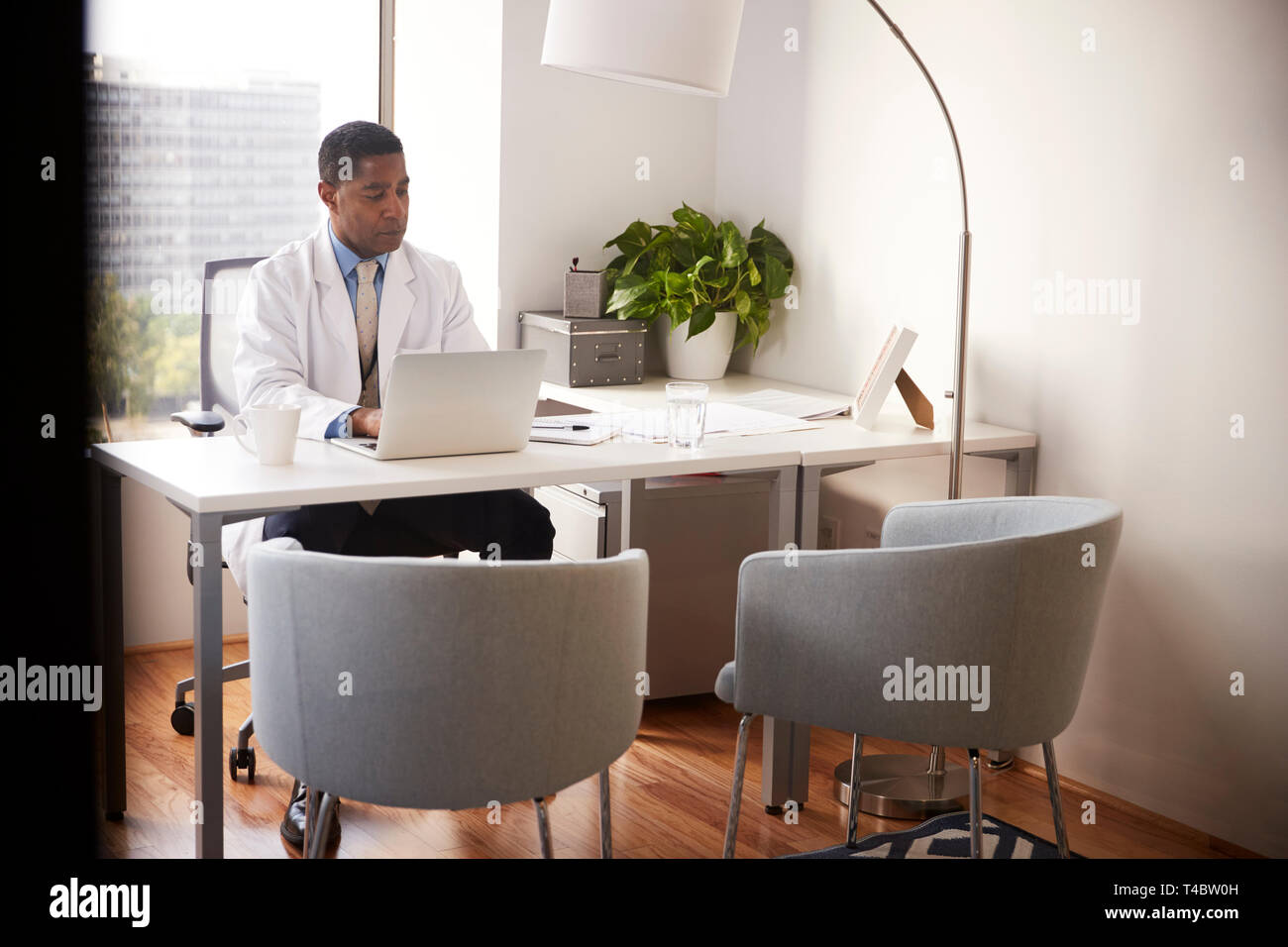 Medico di sesso maschile che indossa mantello bianco in ufficio seduti alla scrivania che lavora sul computer portatile Foto Stock