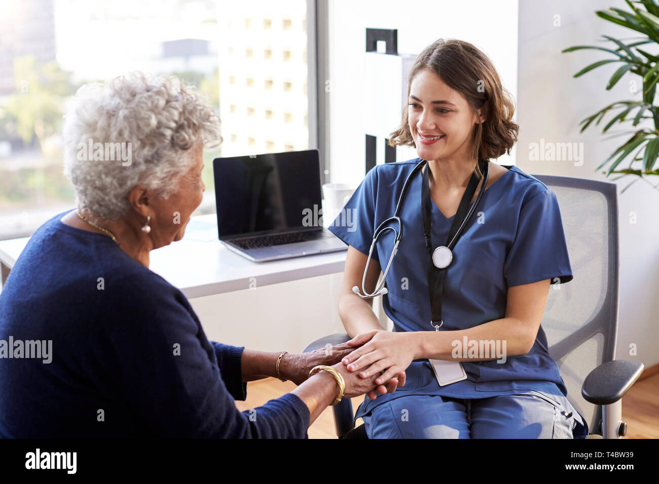 L'infermiera indossando Scrubs In ufficio rassicurante Senior paziente femmina e tenendo le Sue mani Foto Stock
