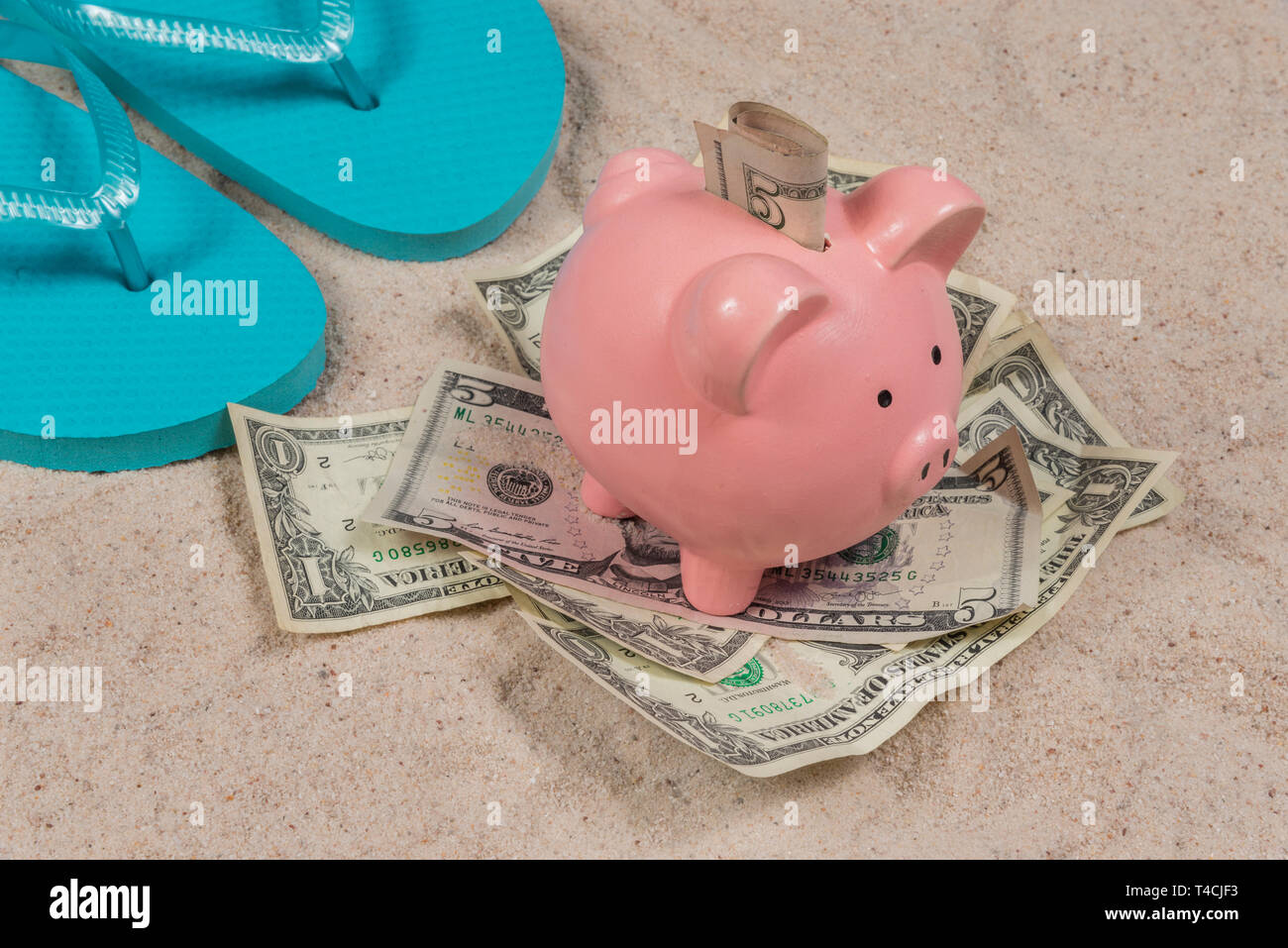 Inquadratura orizzontale di una coppia di blu flip-flop e un rosa salvadanaio su un mucchio di soldi con il denaro nel suo slot su una spiaggia di sabbia. Foto Stock