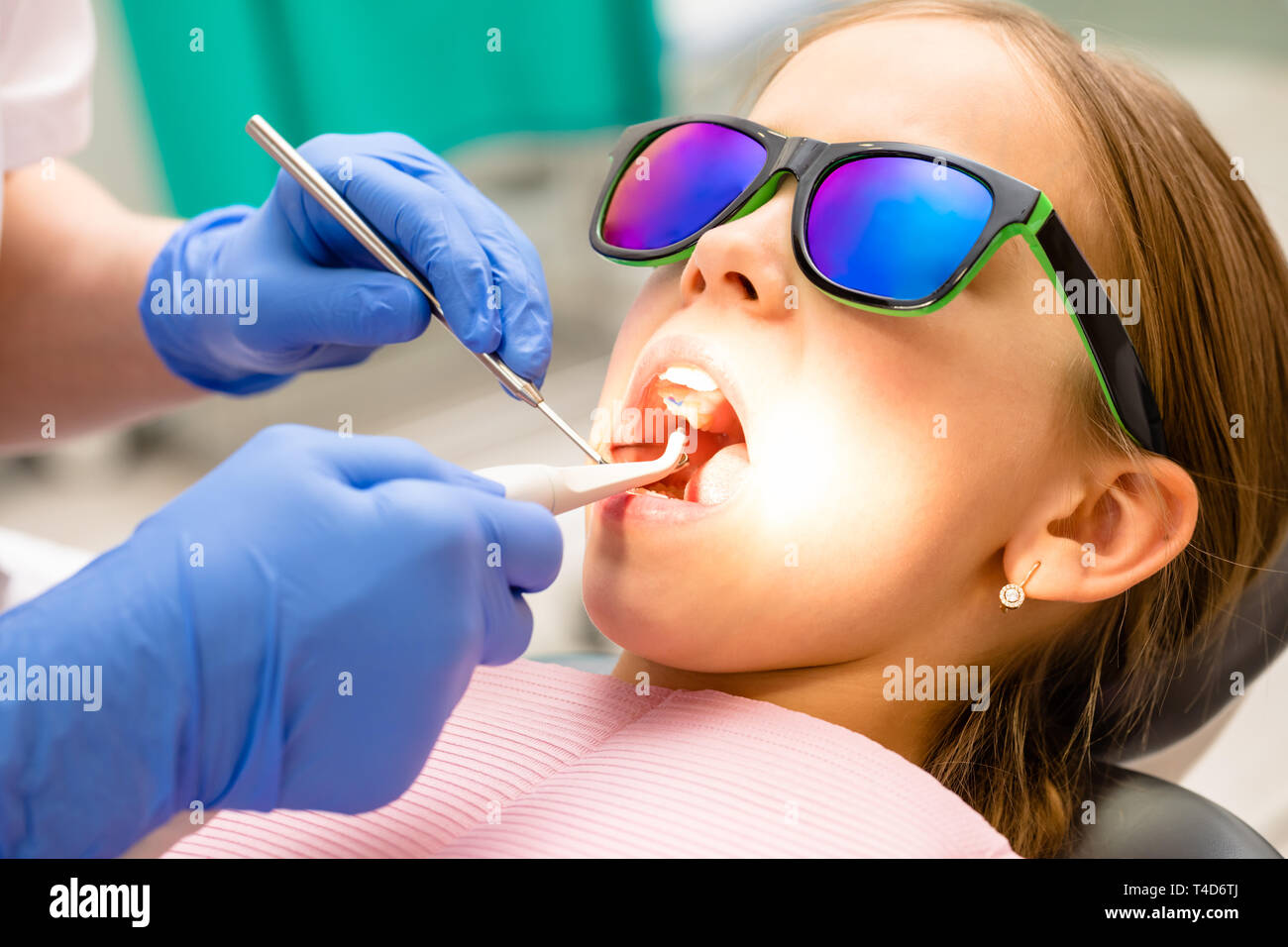 Dentista esaminando denti di elementare età ragazza in pazienti pediatrici clinica dentale utilizzando lo strumento dentale e specchio inclinato. Bambino seduto in una poltrona odontoiatrica indossa Foto Stock