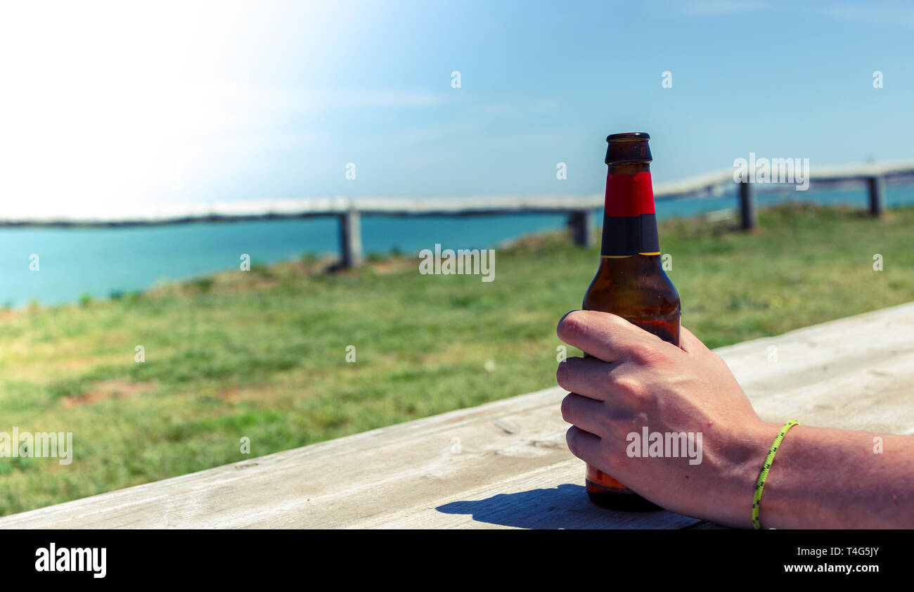 Concetto di vacanza. Maschio di mano che tiene una bottiglia di birra sul mare spiaggia sulla piattaforma di legno Foto Stock