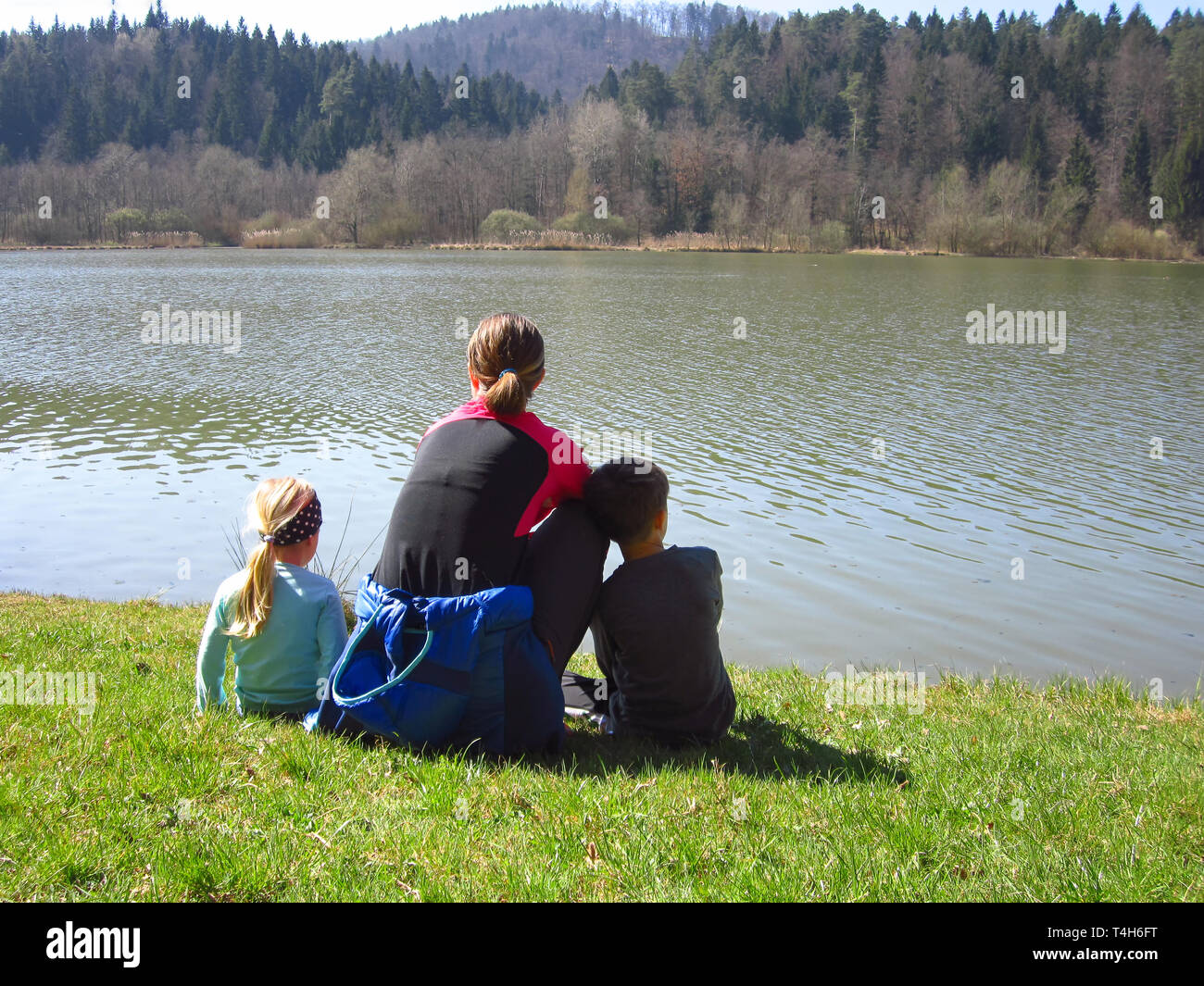Madre e figlio e figlia seduti su un prato di erba verde guardando il lago e la foresta. La famiglia felice in natura Foto Stock