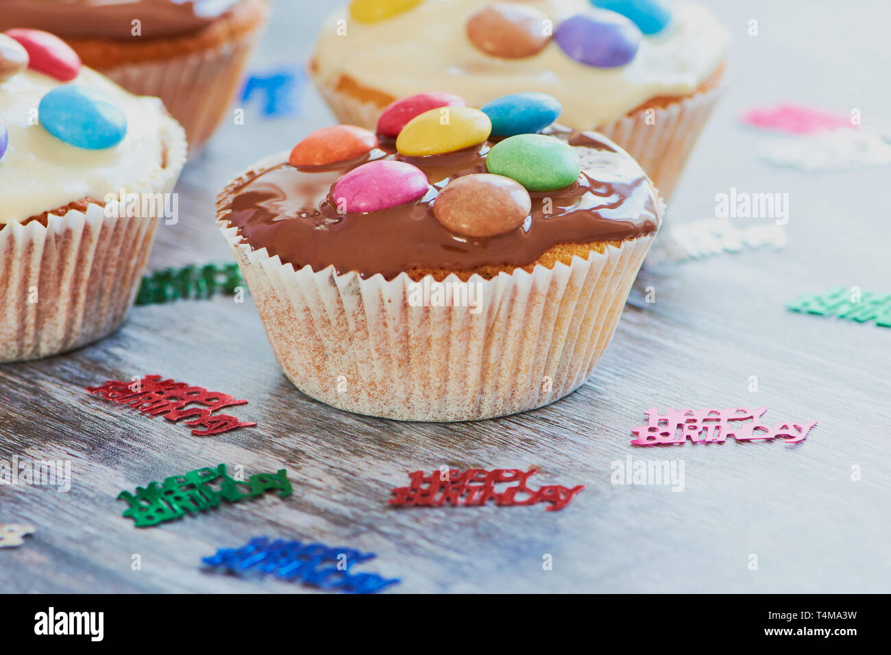 Selfmade muffin rivestiti con cioccolato e lenticchie di cioccolato su una superficie di legno accanto alla festa di compleanno di piccole dimensioni con decor con le parole: Buon compleanno Foto Stock