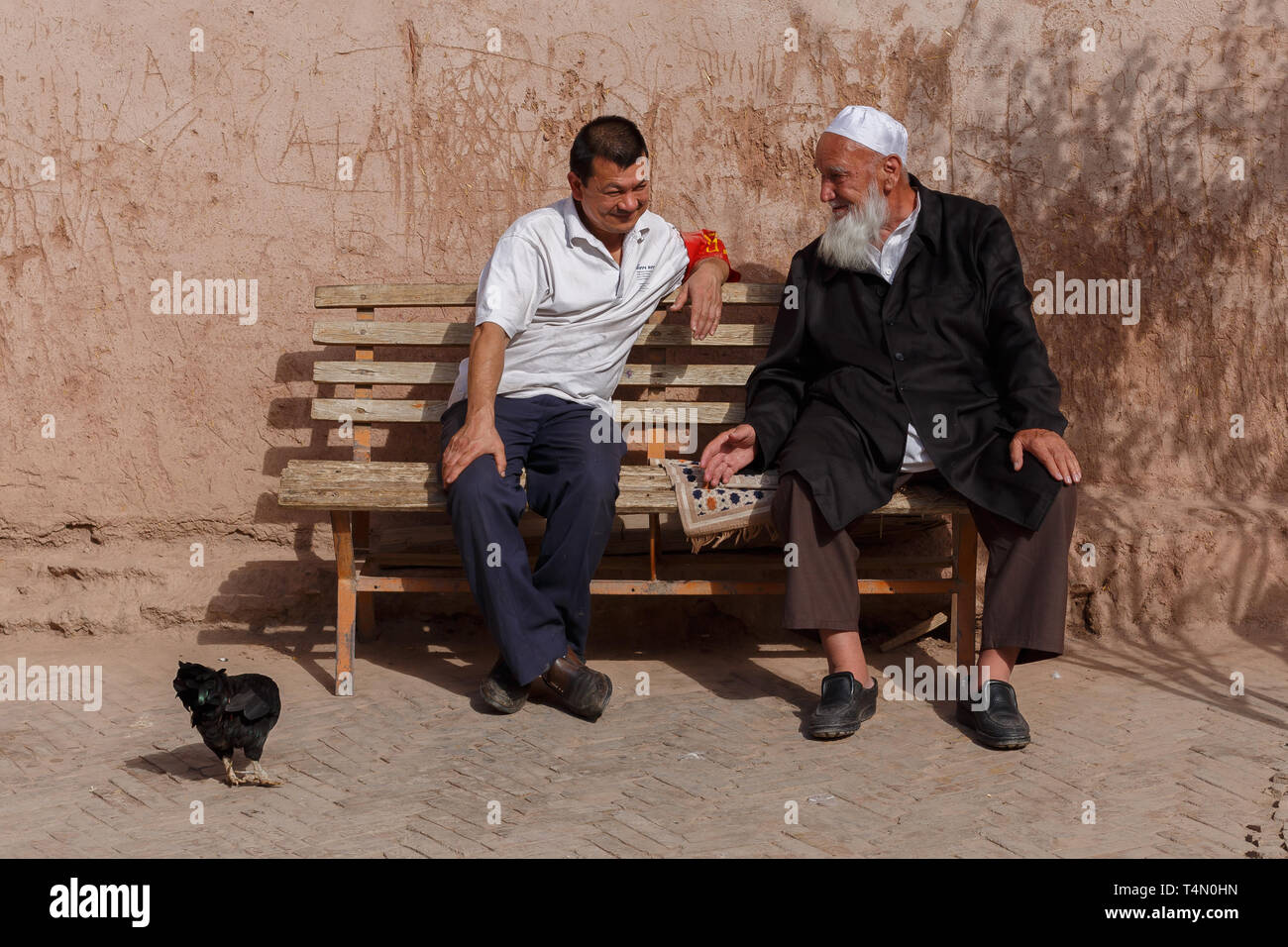 Due uomini uigura avente una conversazione su un banco di lavoro. Notevole dettaglio: il pollo sul lato sinistro dei due colleghi. Kashgar, provincia dello Xinjiang, Cina. Foto Stock