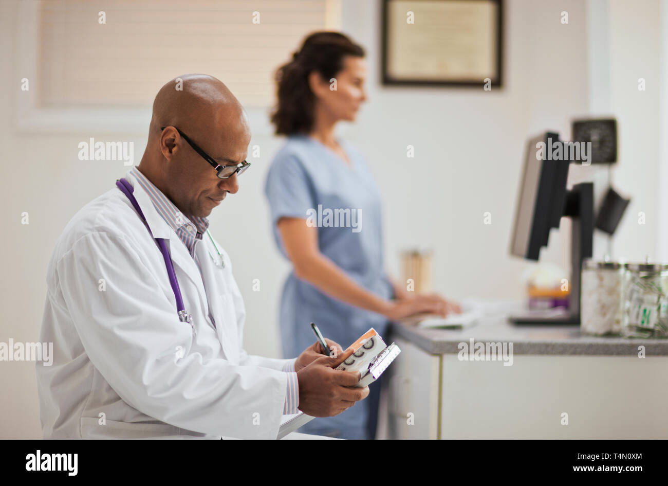 Medico maschio prendendo appunti su appunti come egli tiene una scatola di farmaco mentre nel suo ufficio con un infermiere. Foto Stock