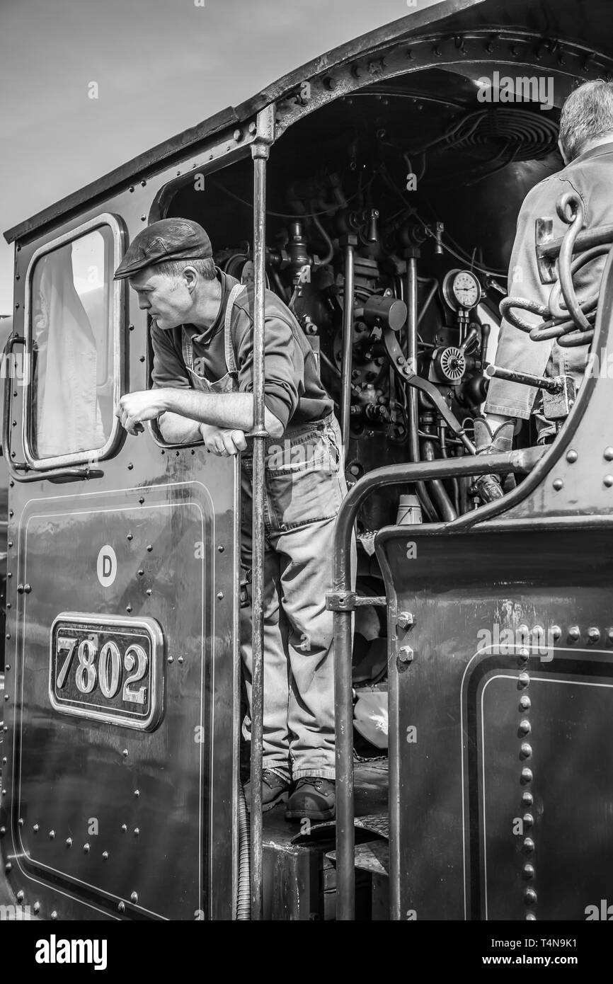 Primo piano in bianco e nero dell'equipaggio del treno, con tute, all'interno della cabina della locomotiva a vapore vintage del Regno Unito. Macchinetta del treno a vapore, in berretto piatto, con vista. Foto Stock