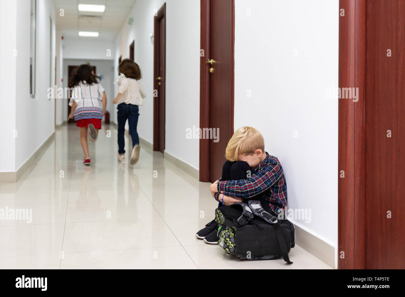 Il pianto piccolo ragazzo seduto sul pavimento della scuola con le mani sulle ginocchia e la testa tra le sue gambe la sofferenza di un atto di bullismo mentre due ragazze scappare Foto Stock