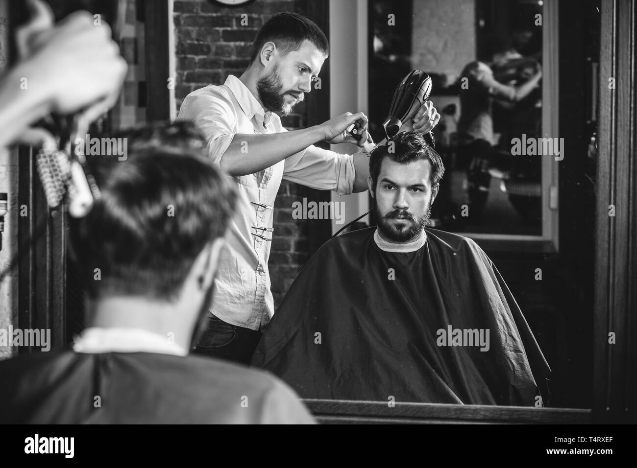 Giovani bel barbiere rendendo haircut per attraente uomo barbuto al barbiere. Nero e bianco o incolore foto. Acconciatura, salone di bellezza e parrucchiere, barbiere, il concetto di stile di vita. Caucasian modelli maschili. Foto Stock