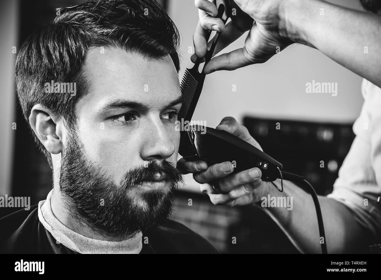 Giovani bel barbiere rendendo haircut per attraente uomo barbuto al barbiere. Nero e bianco o incolore foto. Acconciatura, salone di bellezza e parrucchiere, barbiere, il concetto di stile di vita. Caucasian modelli maschili. Foto Stock