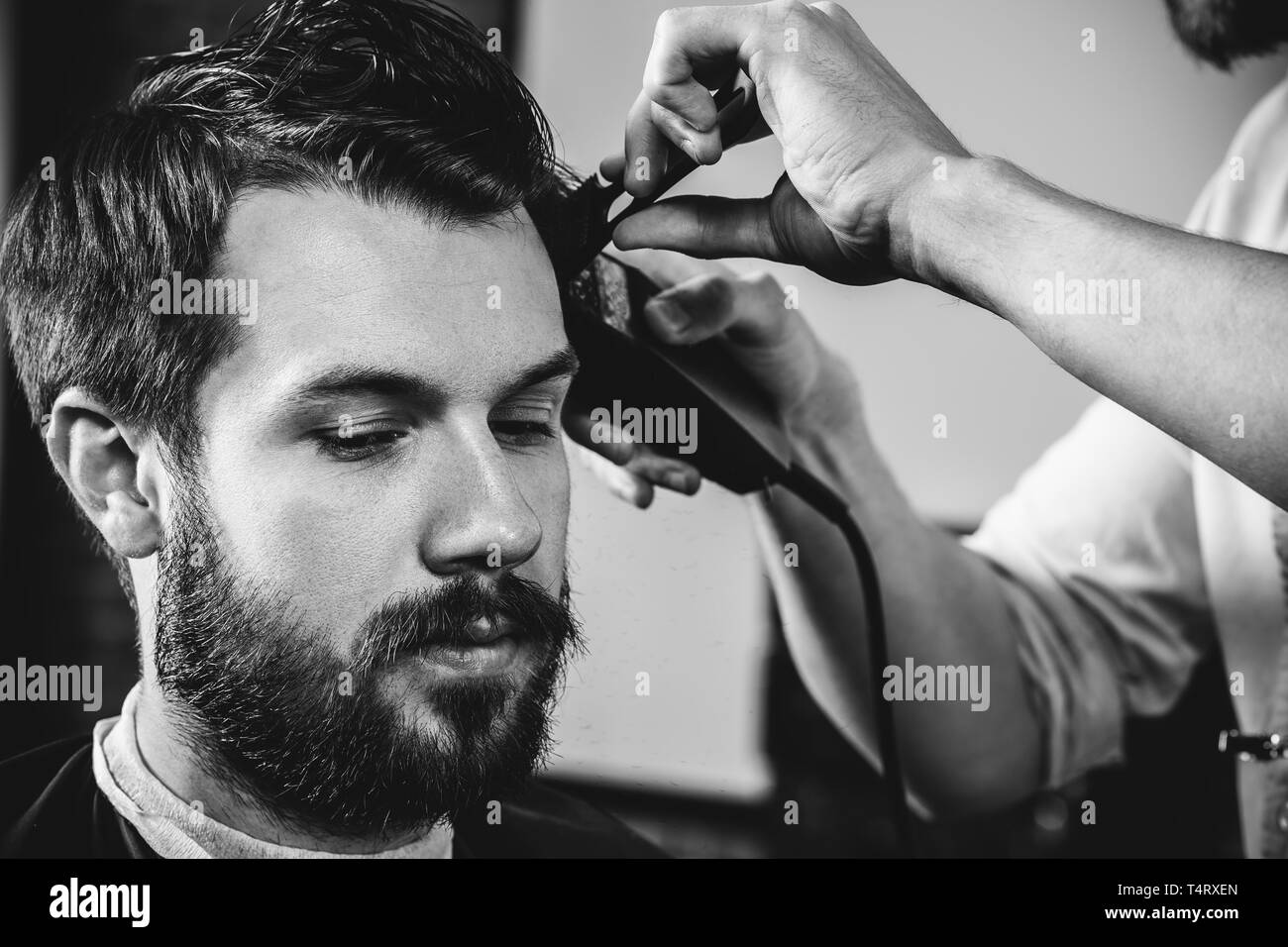 Giovani bel barbiere rendendo haircut per attraente uomo barbuto al barbiere. Nero e bianco o incolore foto. Acconciatura, salone di bellezza e parrucchiere, barbiere, il concetto di stile di vita. Caucasian modelli maschili. Foto Stock