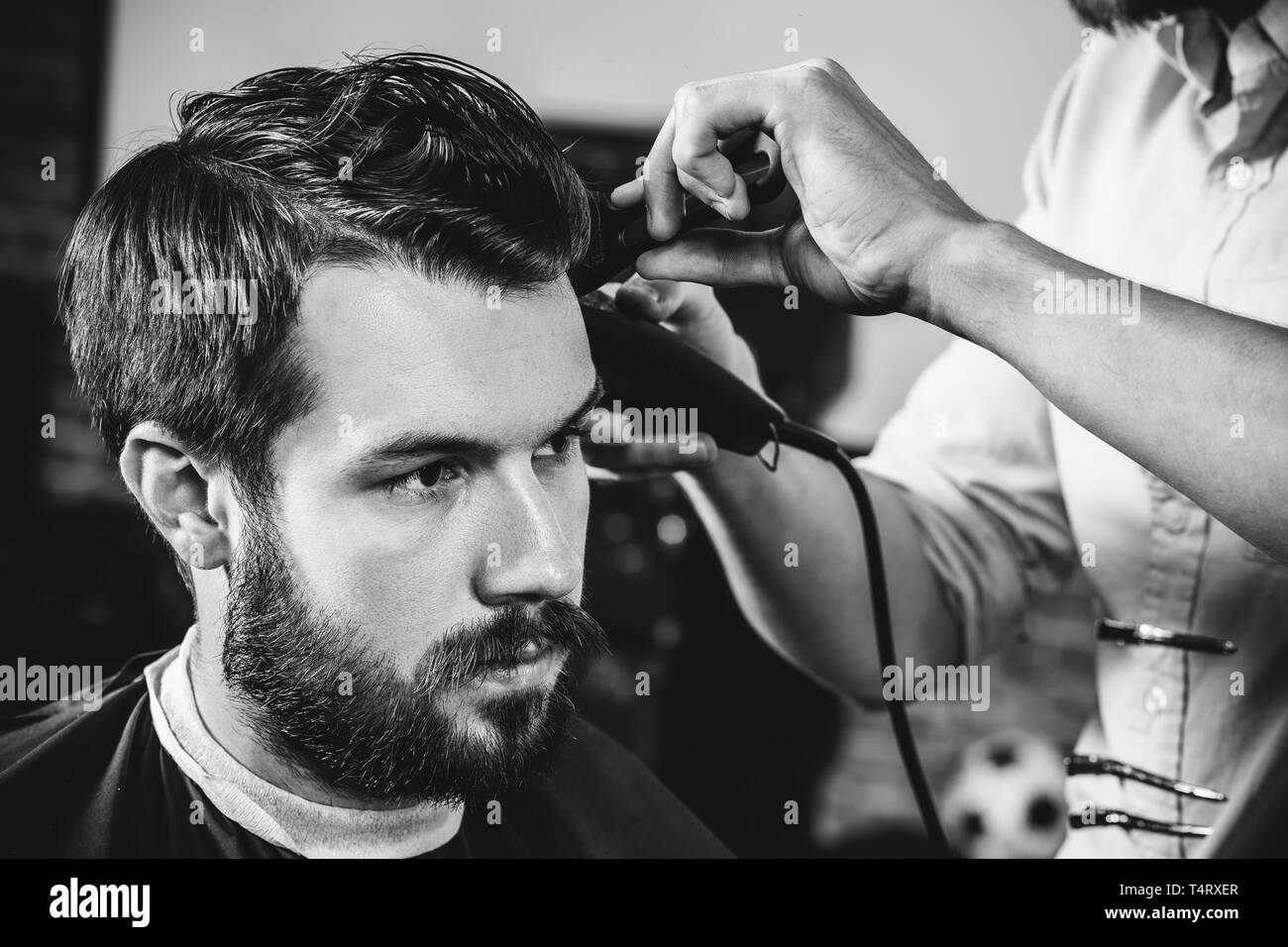 Giovani bel barbiere rendendo haircut per attraente uomo barbuto al barbiere. Nero e bianco o incolore foto. Acconciatura, salone di bellezza e parrucchiere, barbiere, il concetto di stile di vita. Caucasian modelli maschili. Foto Stock
