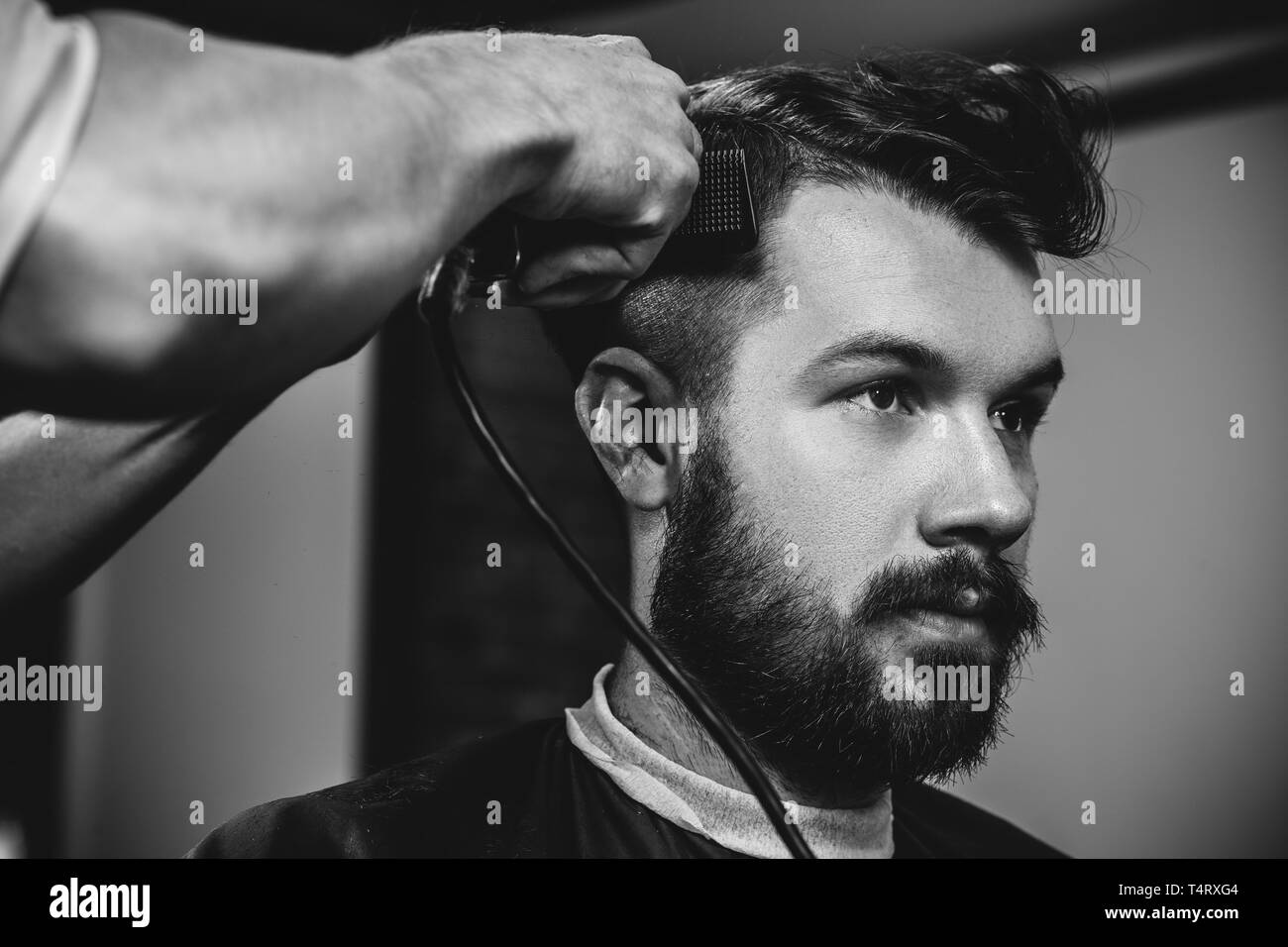 Giovani bel barbiere rendendo haircut per attraente uomo barbuto al barbiere. Nero e bianco o incolore foto. Acconciatura, salone di bellezza e parrucchiere, barbiere, il concetto di stile di vita. Caucasian modelli maschili. Foto Stock