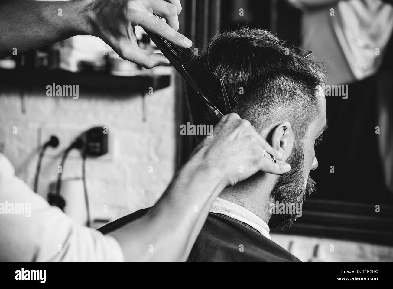 Giovani bel barbiere rendendo haircut per attraente uomo barbuto al barbiere. Nero e bianco o incolore foto. Acconciatura, salone di bellezza e parrucchiere, barbiere, il concetto di stile di vita. Caucasian modelli maschili. Foto Stock