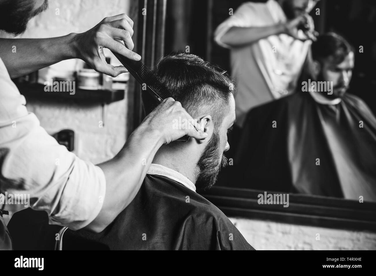 Giovani bel barbiere rendendo haircut per attraente uomo barbuto al barbiere. Nero e bianco o incolore foto. Acconciatura, salone di bellezza e parrucchiere, barbiere, il concetto di stile di vita. Caucasian modelli maschili. Foto Stock
