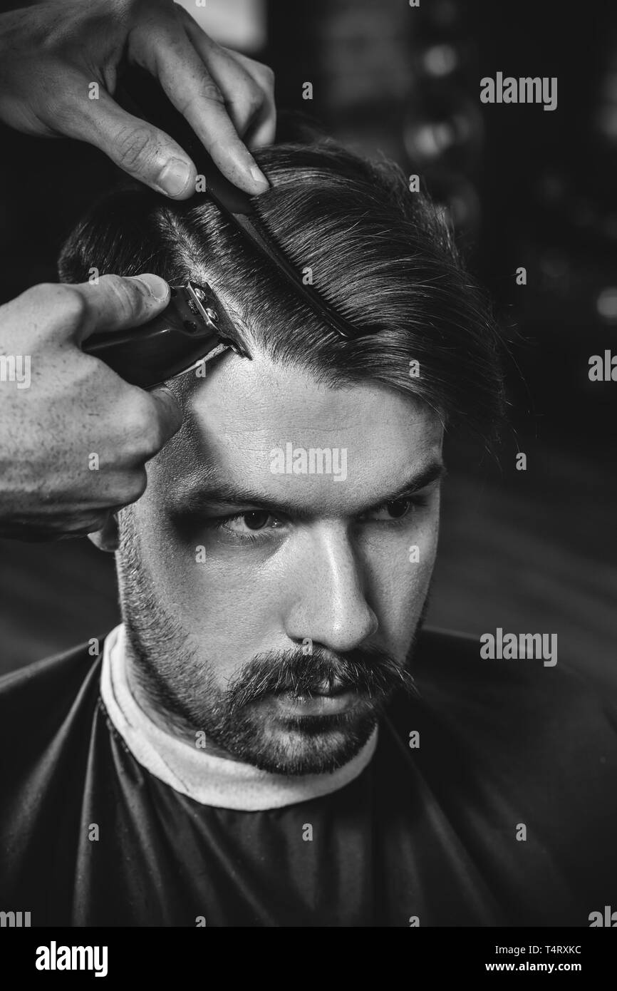 Giovani bel barbiere rendendo haircut per attraente uomo barbuto al barbiere. Nero e bianco o incolore foto. Acconciatura, salone di bellezza e parrucchiere, barbiere, il concetto di stile di vita. Caucasian modelli maschili. Foto Stock