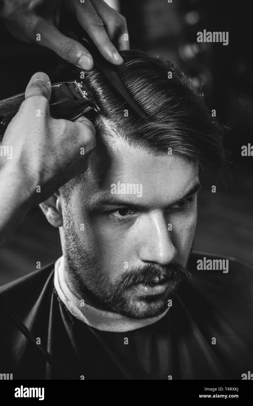 Giovani bel barbiere rendendo haircut per attraente uomo barbuto al barbiere. Nero e bianco o incolore foto. Acconciatura, salone di bellezza e parrucchiere, barbiere, il concetto di stile di vita. Caucasian modelli maschili. Foto Stock