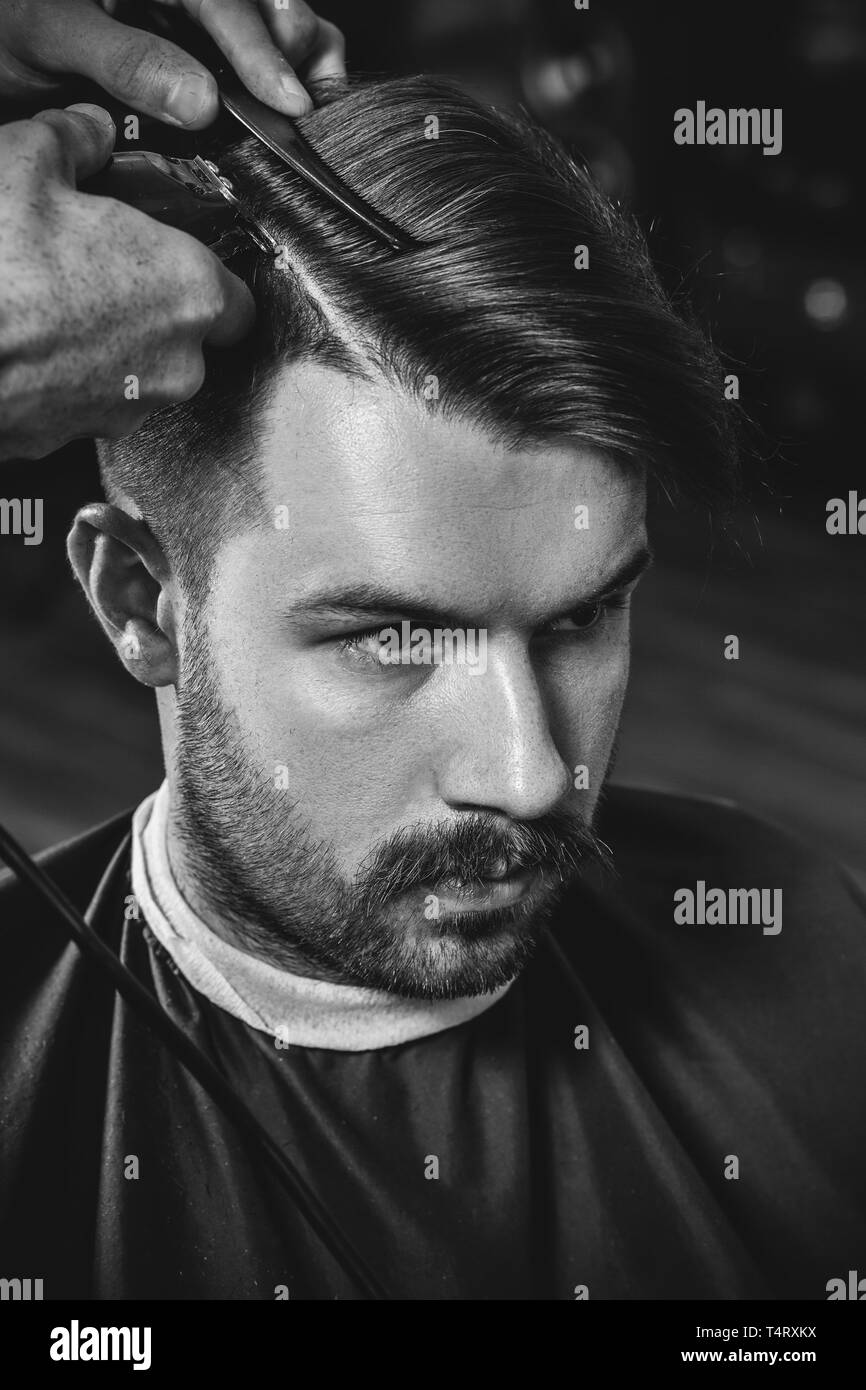 Giovani bel barbiere rendendo haircut per attraente uomo barbuto al barbiere. Nero e bianco o incolore foto. Acconciatura, salone di bellezza e parrucchiere, barbiere, il concetto di stile di vita. Caucasian modelli maschili. Foto Stock
