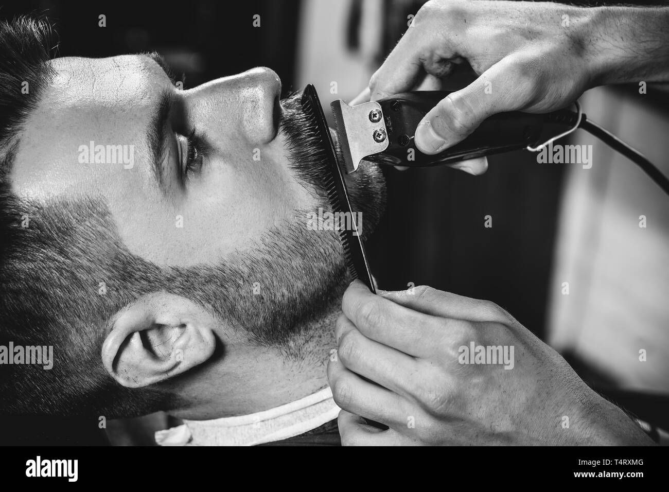 Client durante la rasatura della barba in barberia. Giovani bel barbiere e attraente uomo barbuto. Nero e bianco o incolore foto. Acconciatura, salone di bellezza e parrucchiere, barbiere, il concetto di stile di vita. Caucasian modelli maschili. Foto Stock