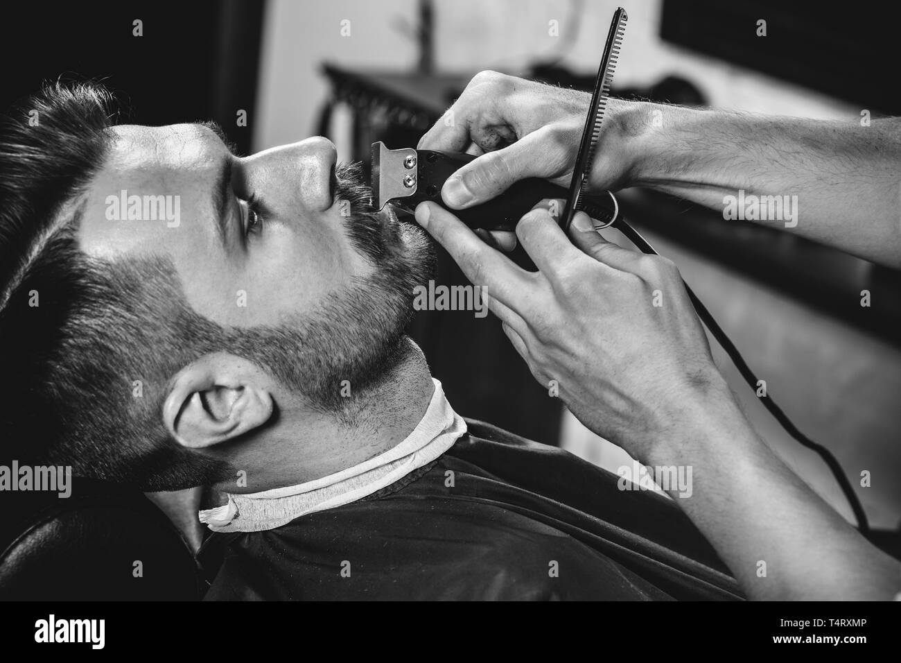Client durante la rasatura della barba in barberia. Giovani bel barbiere e attraente uomo barbuto. Nero e bianco o incolore foto. Acconciatura, salone di bellezza e parrucchiere, barbiere, il concetto di stile di vita. Caucasian modelli maschili. Foto Stock
